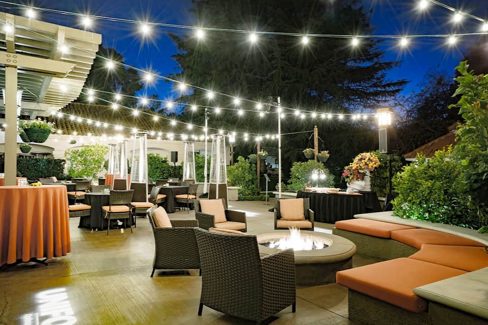 Elegant outdoor event space with string lights, fire pit, orange linens, and modern seating at dusk