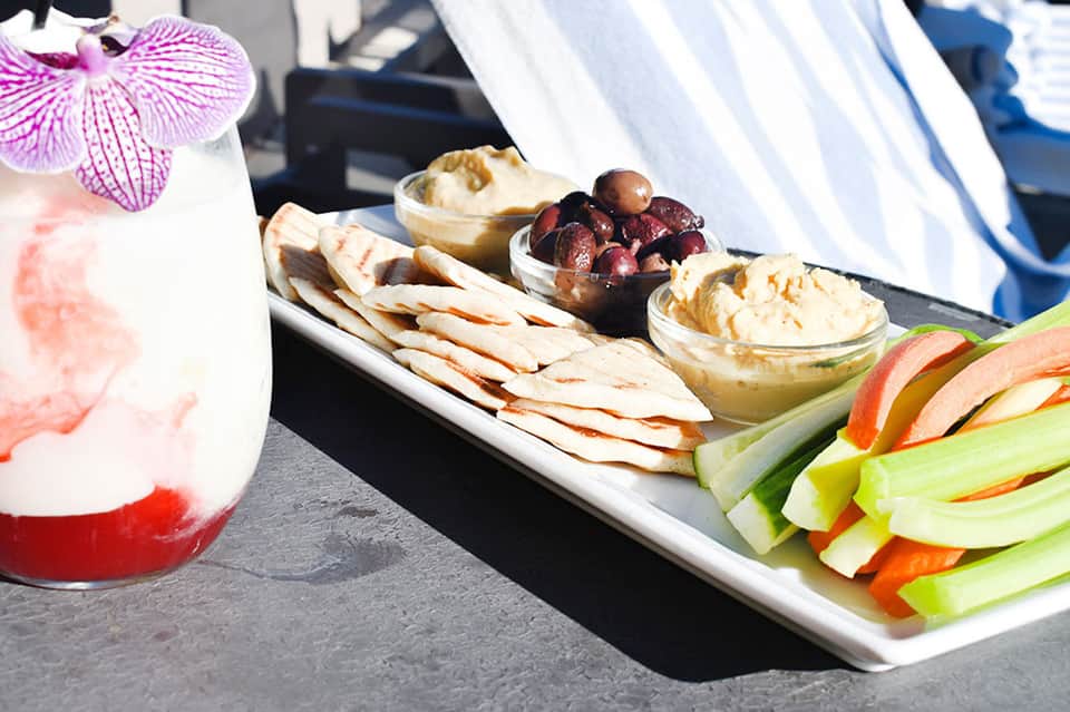 Poolside appetizer platter with pink cocktail, flatbread, dips, olives, and fresh vegetables on white plate