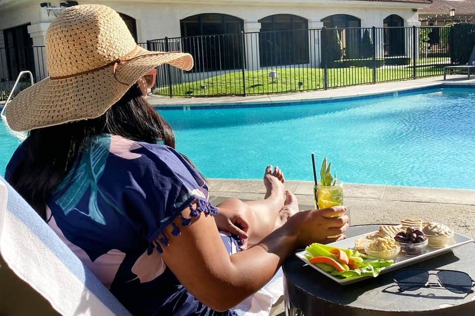 Woman relaxing poolside wearing straw hat with beverage, food plate, and fenced pool in background
