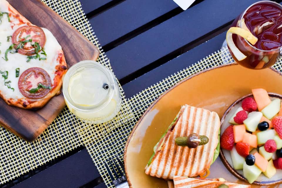 Poolside food spread: gourmet pizza, charcuterie sandwich, fresh fruit salad, and white wine on striped lounge chair