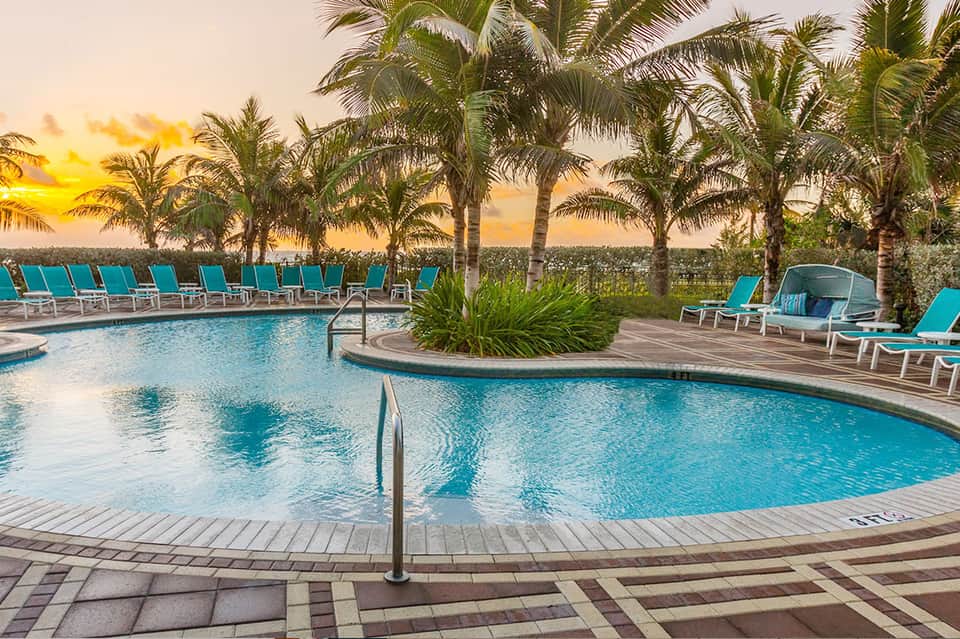 Resort pool at sunset with turquoise water, palm trees, lounge chairs, and colorful sky