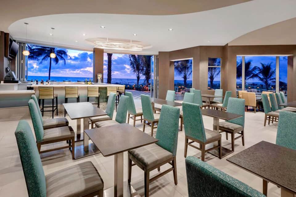 Upscale beachfront dining room with turquoise chairs, tables, and panoramic ocean views at dusk