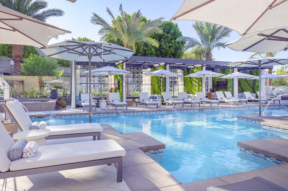 Luxury resort pool with blue water, white lounge chairs, large umbrellas, and palm trees under clear sky