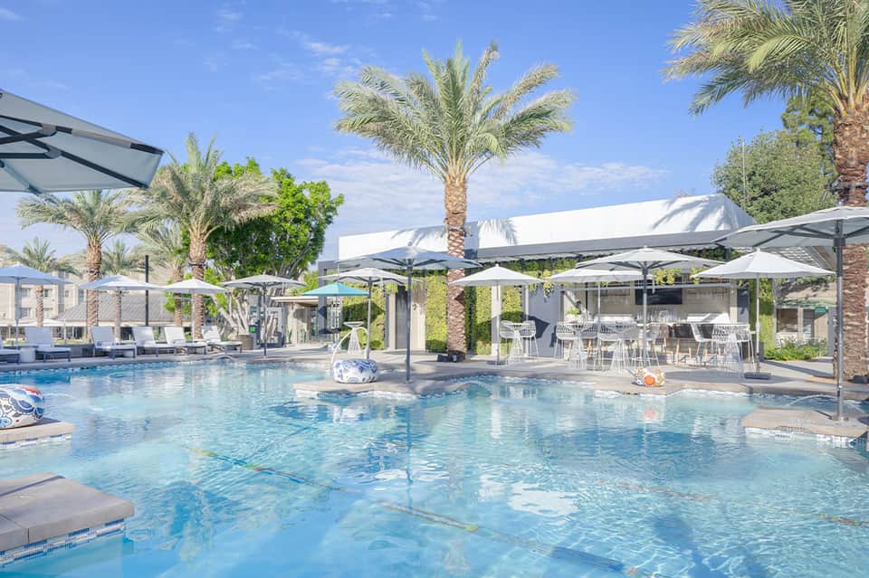 Bright pool area with palm trees, white umbrellas, modern loungers, and white resort buildings under blue sky