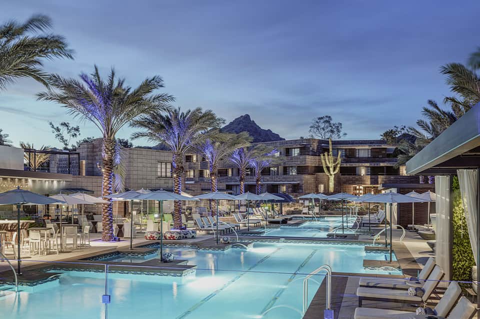 Resort pool area at night with illuminated palm trees, lounge chairs, umbrellas, and mountain backdrop