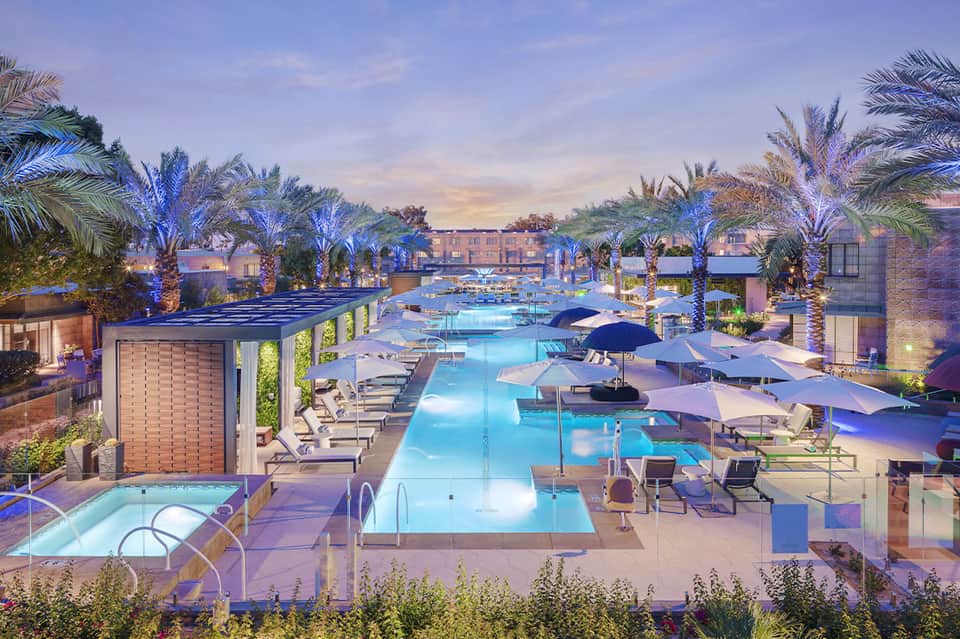 Illuminated resort pool complex at dusk with palm trees and blue-lit umbrellas