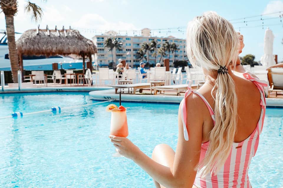 Woman in pink swimsuit holding a cocktail by the pool with resort buildings and ocean view