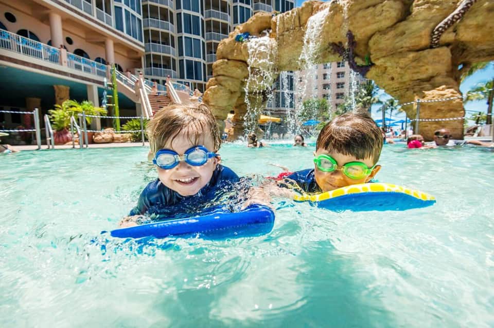 Two young children swimming with kickboards in a resort pool beneath large rock formations