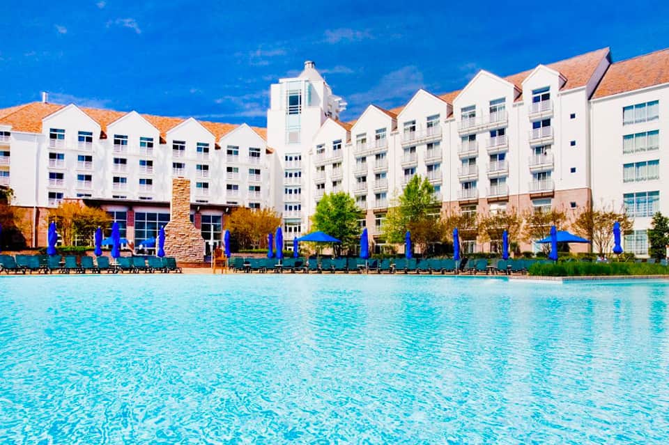 Large resort with white multi-story buildings, terracotta roofs, and crystal-clear turquoise pool with blue umbrellas