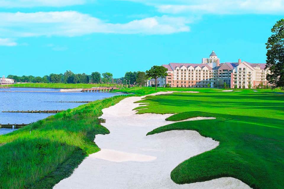 Luxury waterfront resort with manicured golf course, sand bunkers, and elegant colonial-style architecture overlooking a bay.