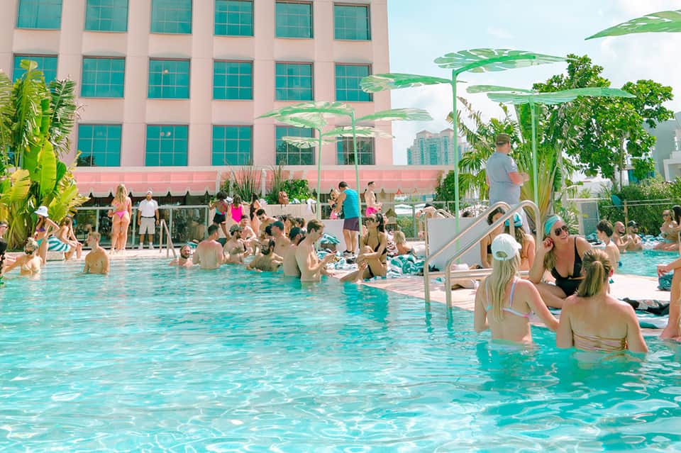 Busy outdoor pool area with guests swimming and lounging under green umbrellas