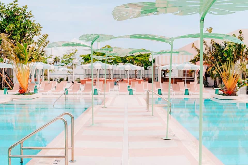 Outdoor resort pool with turquoise water, green pergola shade structures, pink loungers, and tropical plants