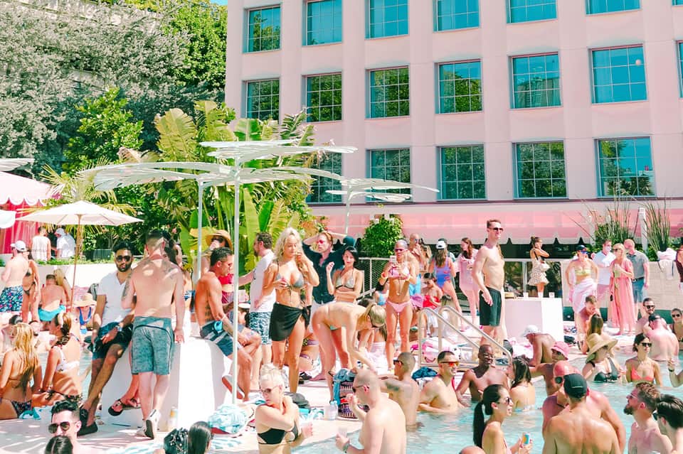 Crowded pink-themed resort pool party with guests in water, umbrellas, and modern building in background