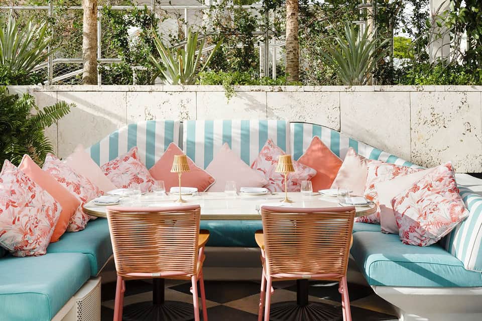 Outdoor dining area with turquoise striped banquette seating, coral and pink patterned pillows, and tropical garden backdrop