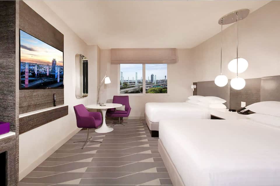 Modern hotel bedroom with two beds, city views, purple chairs, and pendant lighting