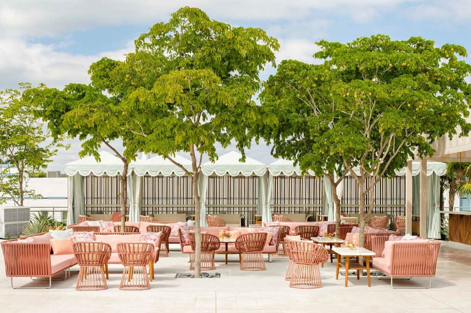 Upscale outdoor terrace with coral lounge seating, shade structures, and mature trees overlooking water