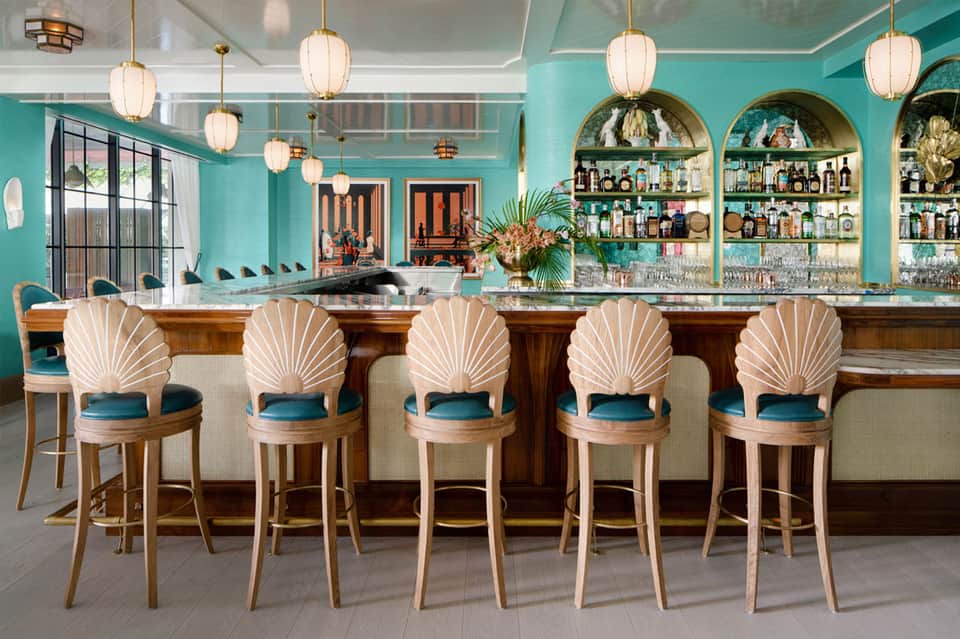 Upscale bar with turquoise walls, shell-backed bar stools, brass pendant lights, and illuminated bottle shelves