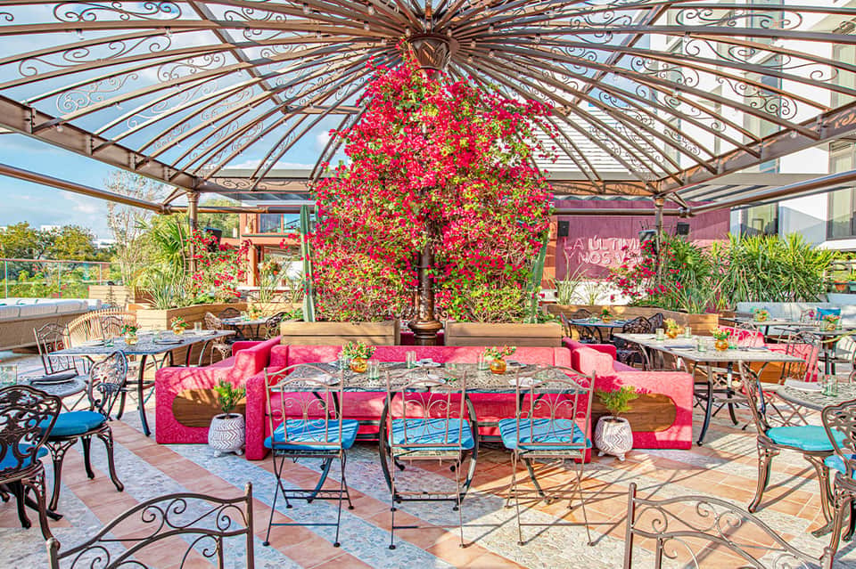 Vibrant outdoor terrace with pink bougainvillea, ornate iron pergola, pink seating, and decorative blue chairs