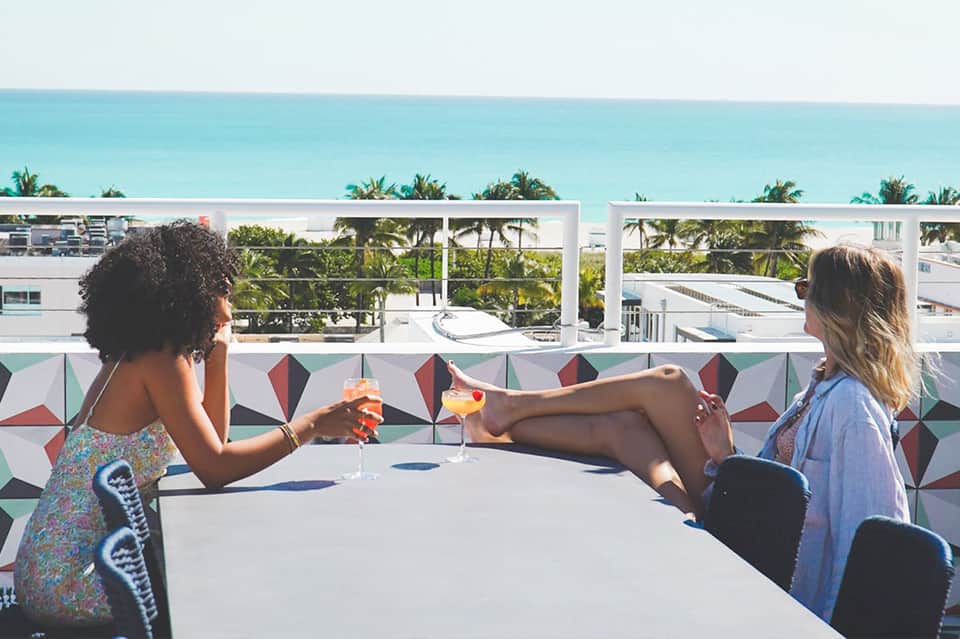 Two women clinking colorful cocktails on a rooftop terrace overlooking palm trees and ocean