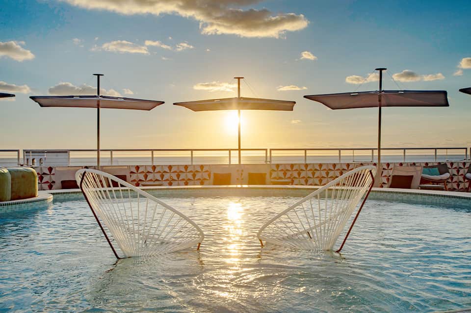 Rooftop pool at sunset with white lounge chairs, umbrellas, and ocean view beyond railings