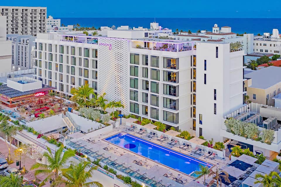 Beachfront hotel exterior with white building, blue pool, palm trees, and ocean view