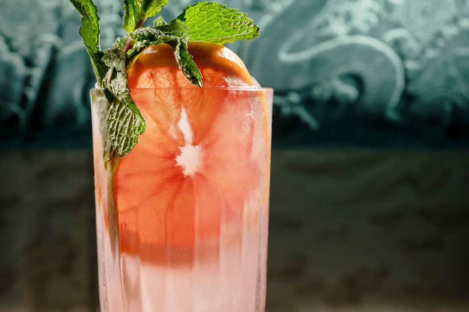 Coral-colored cocktail with mint garnish and fresh fruit in glass on dark background