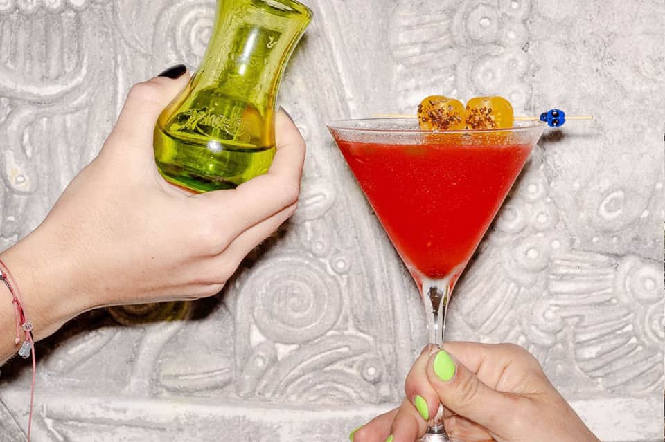 Hand holding green bottle and red cocktail with fruit garnish on textured surface