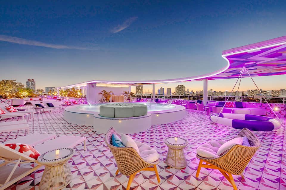 Rooftop lounge with pink neon lighting, modern furniture, and city skyline at dusk