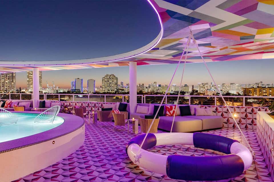 Modern rooftop pool lounge with purple lighting, city skyline view, and seating areas