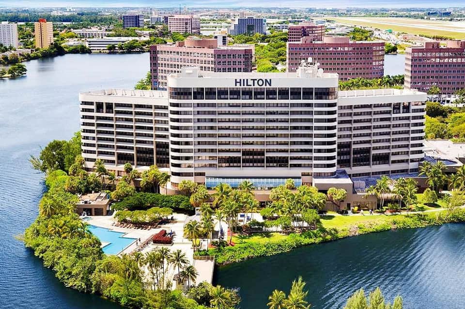 Aerial view of Hilton hotel on waterfront with pool, palm trees, and surrounding cityscape