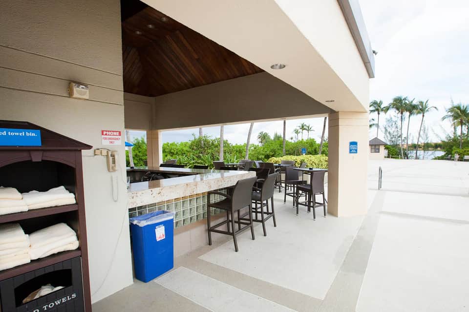 Covered poolside bar area with stone counter, black bar stools, towel bins, and tropical views