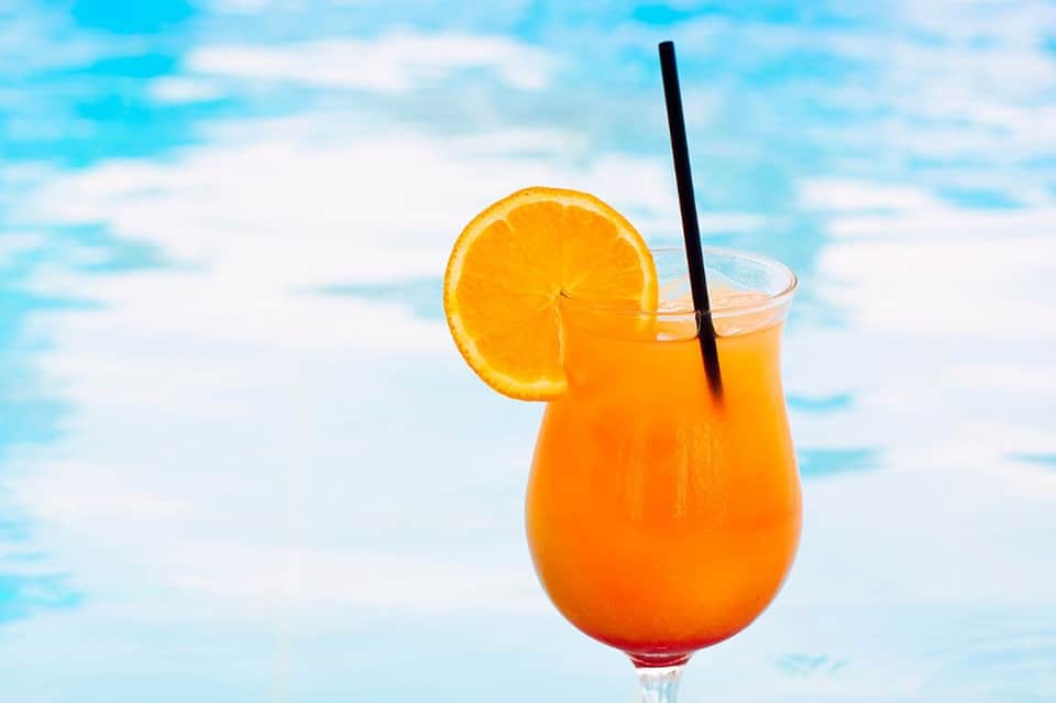 Orange cocktail with orange slice garnish and black straw by poolside