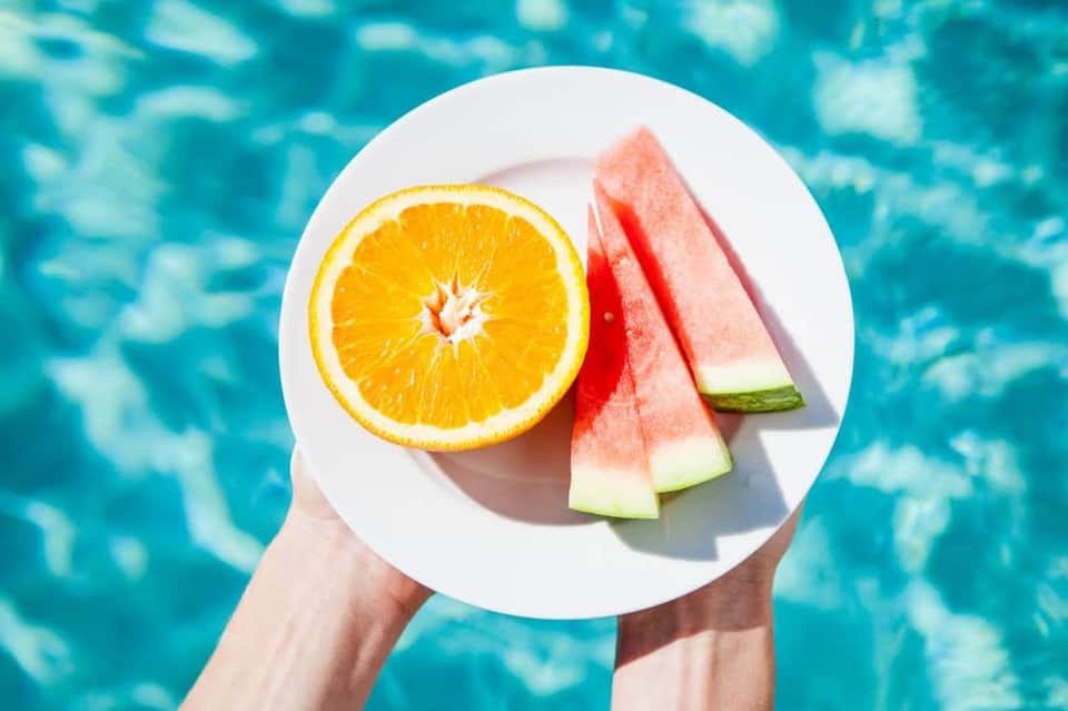 Hands holding plate of fresh fruit including orange slice and watermelon above sparkling pool water