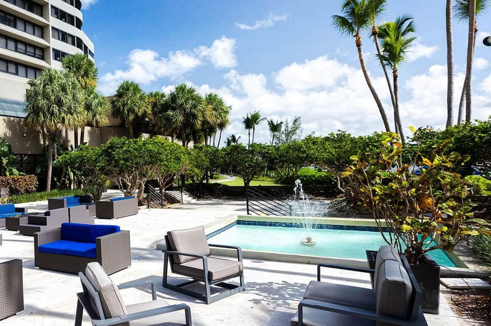 Resort pool area with palm landscaping, modern lounge furniture, and fountain feature
