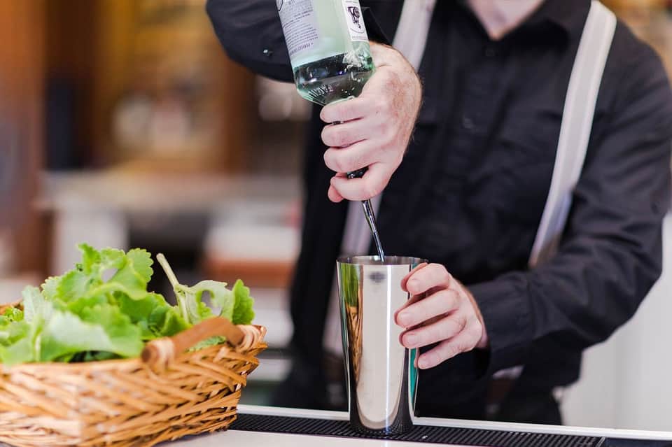 Bartender pouring clear cocktail into glass with fresh vegetables nearby