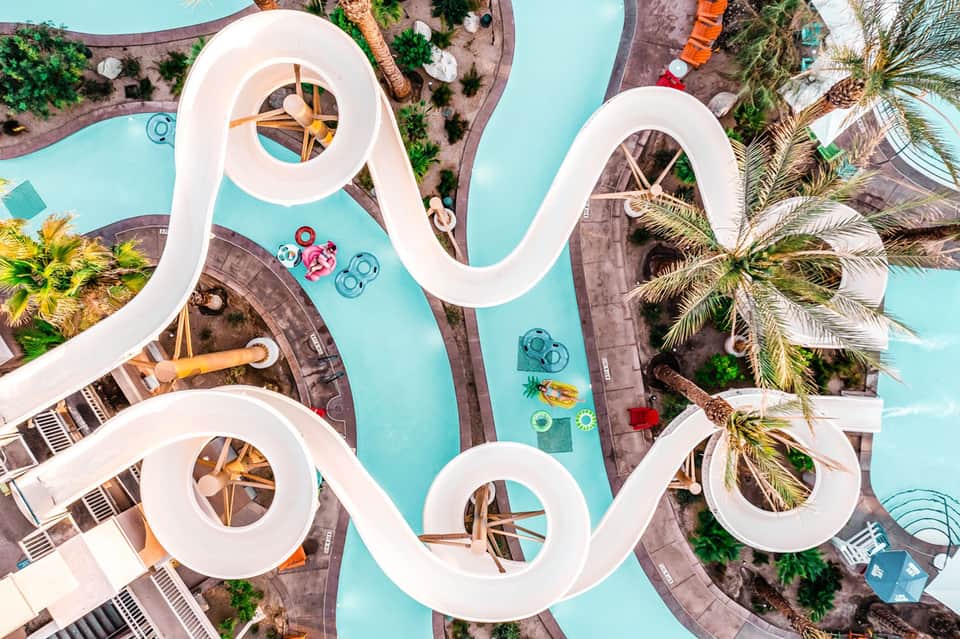 Aerial view of white water slide and turquoise pool with lounge areas and palm trees