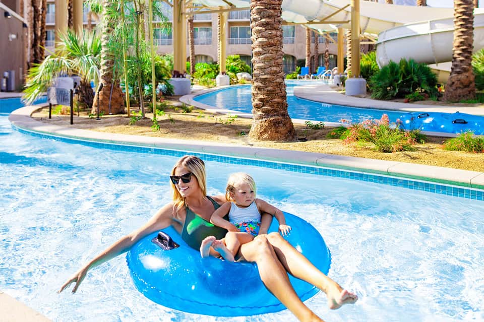 Woman and child enjoying a lazy river on blue float at resort with palm trees and pools