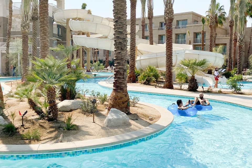 Resort pool area with palm trees, white umbrellas, lounge chairs, and multi-level tan buildings in background
