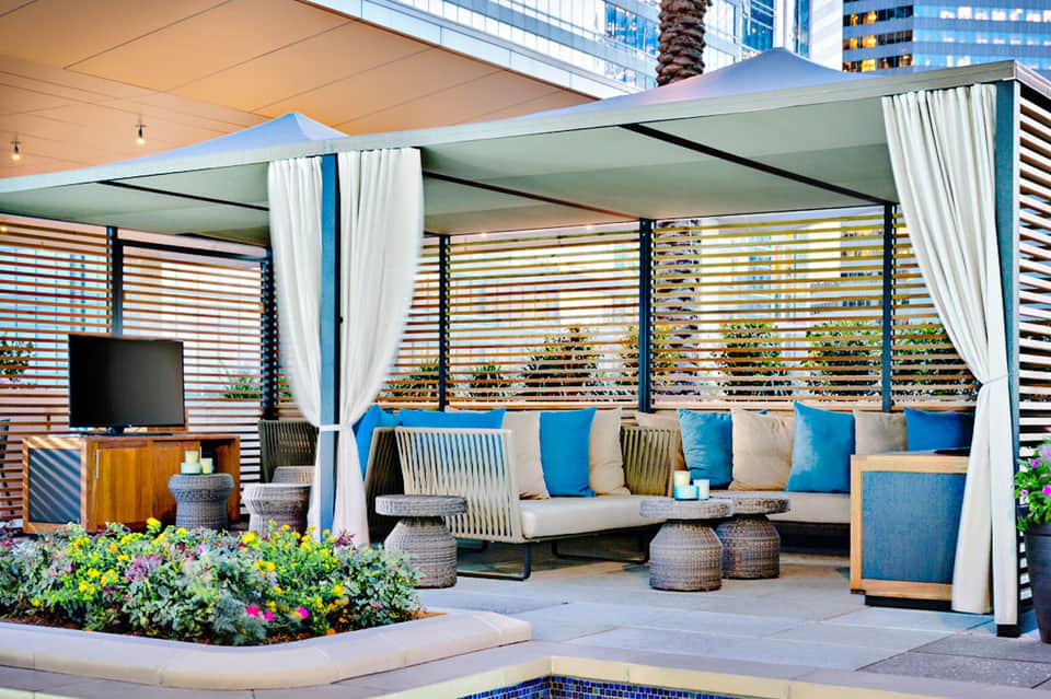 Modern poolside lounge area with blue and white seating, striped privacy screens, white curtains, and potted flowers