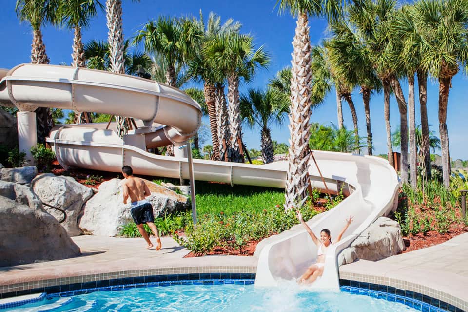 Family water slide at resort pool surrounded by palm trees with children playing