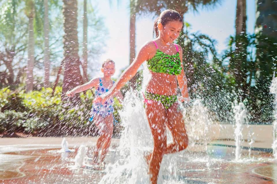 Children playing in outdoor water splash pad surrounded by palm trees on sunny day