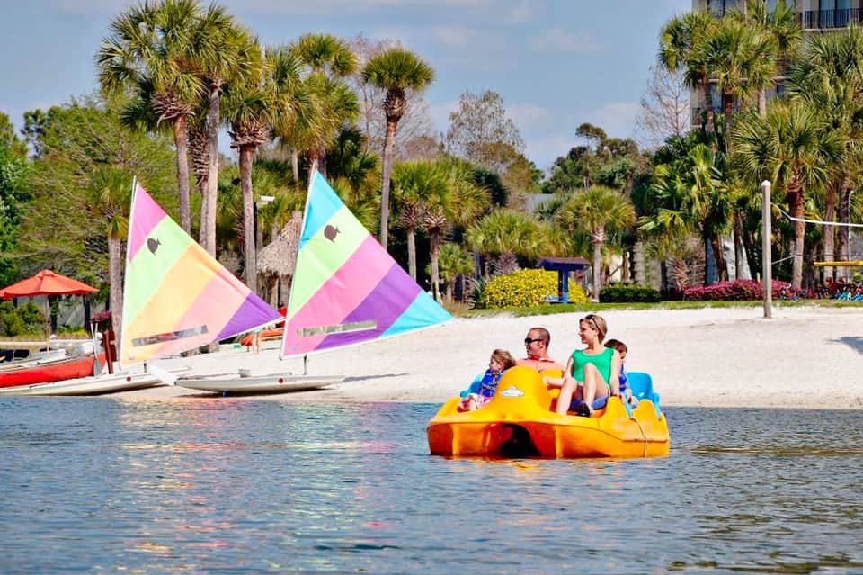 Family enjoying bright yellow paddle boat on beach with colorful sailboats and palm trees