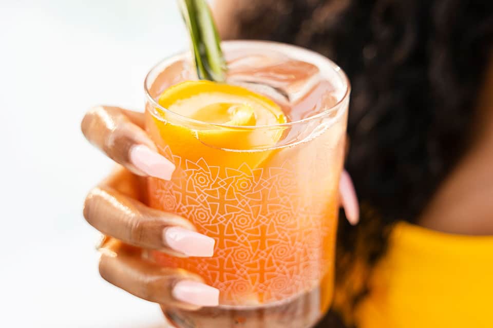 Person holding an orange tropical cocktail with lemon slice and decorative patterned glass