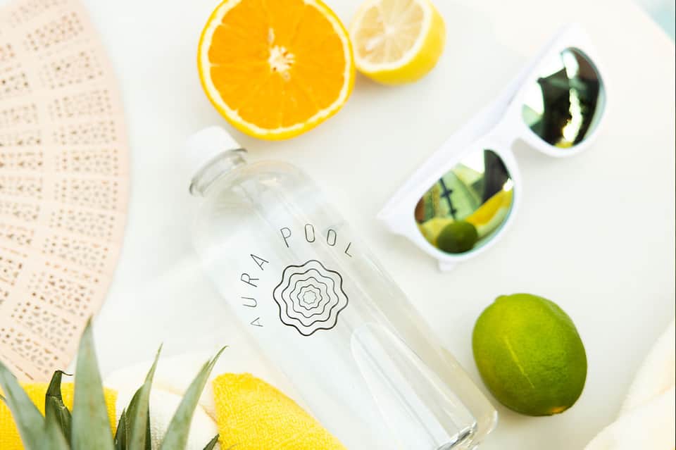 Flat lay of water bottle labeled 'Aura Pool', citrus fruits, sunglasses, and beach accessories on white surface