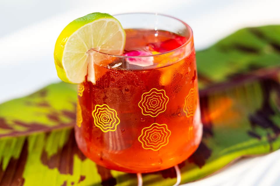 Red cocktail with lime slice and geometric pattern glass garnish against blurred green foliage