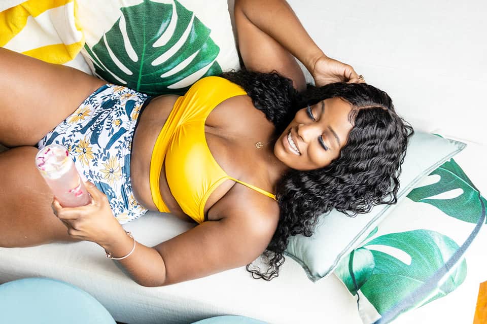 Woman in yellow bikini relaxing on white cushions with tropical leaf pillows and refreshing drink