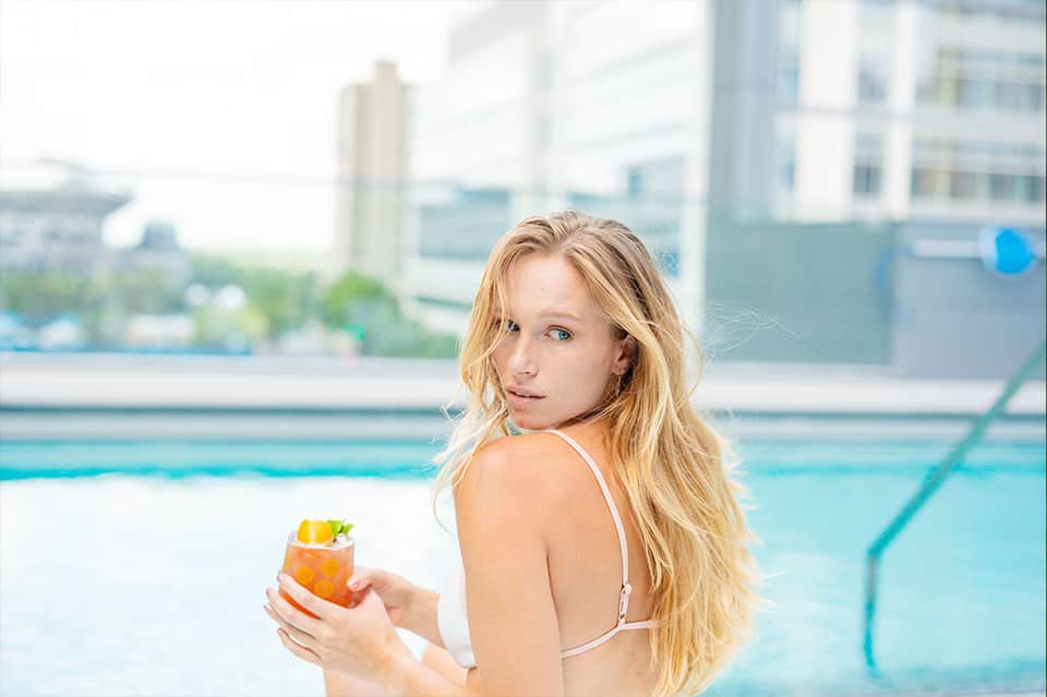 Woman in bikini holding cocktail by hotel rooftop pool with city skyline view