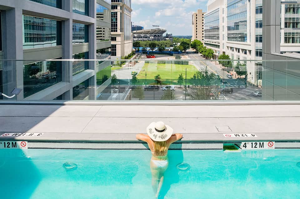 Woman in white hat floating in bright turquoise rooftop pool overlooking urban cityscape