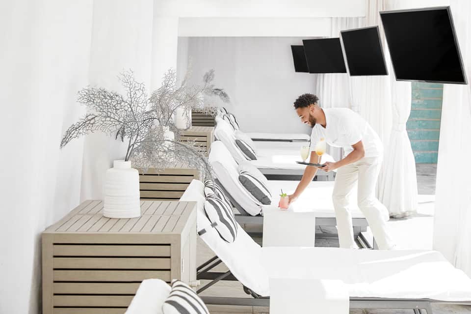 Modern spa lounge with white loungers, potted plants, and staff member serving beverages