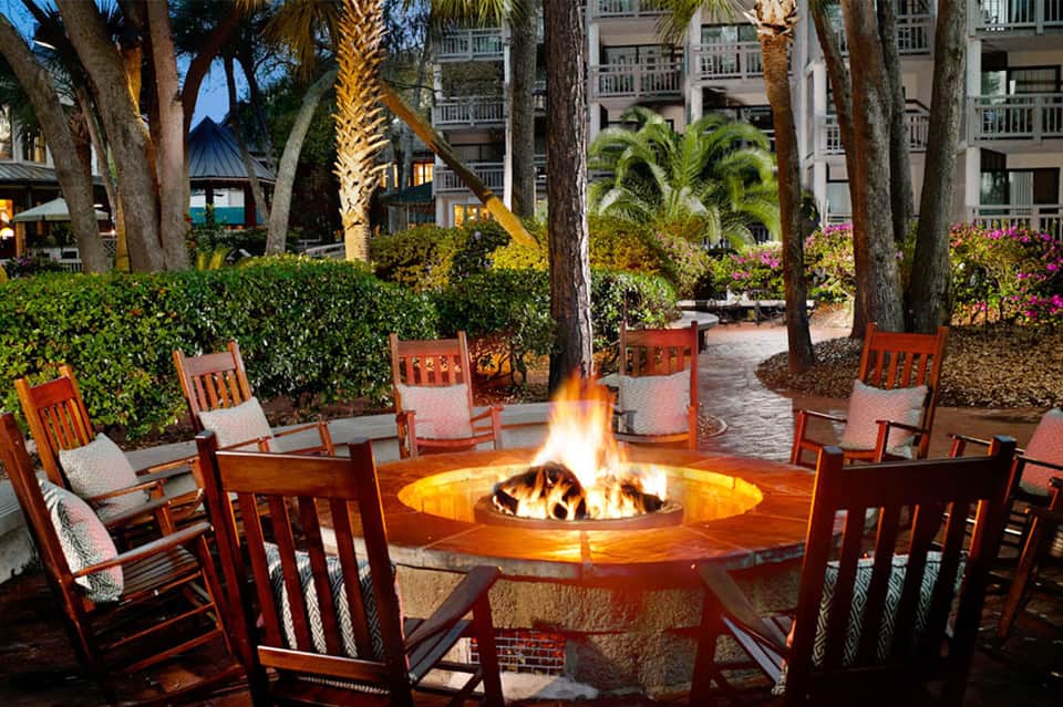 Outdoor fire pit surrounded by rocking chairs under palm trees at beachfront resort